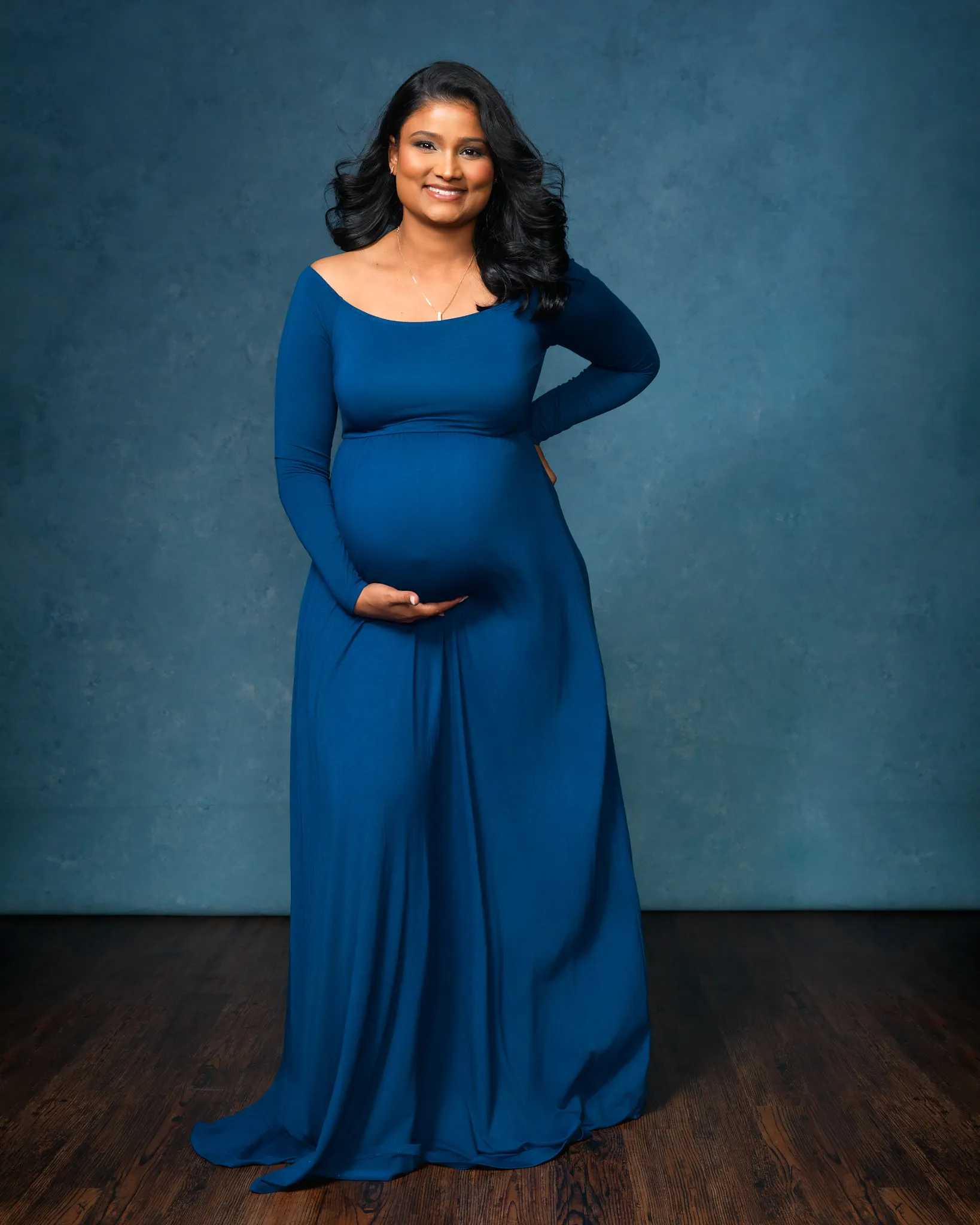 Darkly lit maternity photo of indian woman wearing a blue dress posed in front of a dark blue backdrop