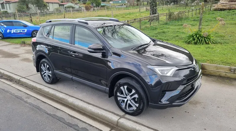 A black Toyota RAV4 SUV is parked on the side of a suburban road. The vehicle features silver alloy wheels and a sleek design. In the background, there are green fields, trees, and a few houses under overcast skies. A blue car with 