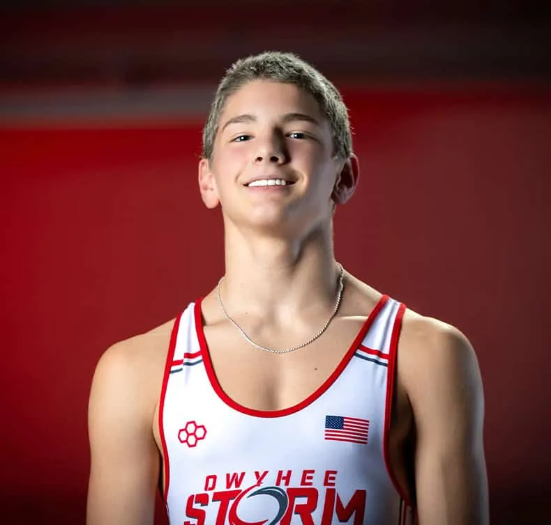  Young Owyhee Storm high school wrestler smiling confidently in white and red USA singlet with American flag patch, wearing silver chain necklace, photographed against red background for professional sports team portrait