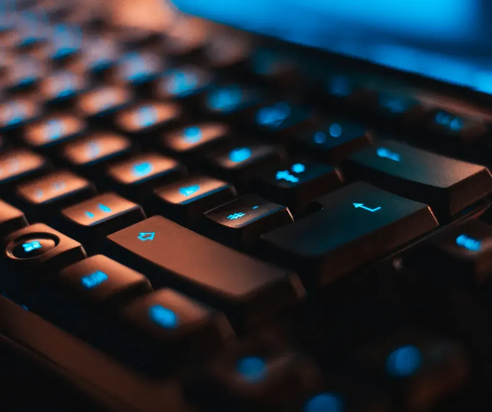 A close-up view of a computer keyboard, illuminated by blue and orange lighting, highlighting the keys with a soft glow. The image focuses on the central part of the keyboard, creating a visually appealing contrast between the dark keys and the vibrant lighting.