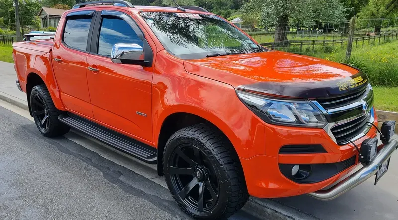 A bright orange Holden Colorado pickup truck is parked on the side of a suburban road. The vehicle features black alloy wheels, chrome side mirrors, a bull bar with mounted spotlights, and tinted windows. The truck has branding on the bonnet reading 