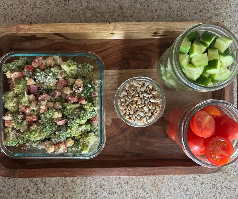 Meal-prep containers with broccoli quinoa salad, chopped cucumbers, tomatoes, and sunflower seeds ready for easy lunches throughout the week.