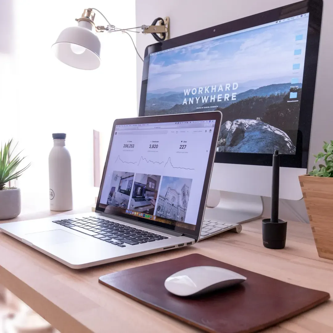 A well-organized workspace with a laptop displaying graphs and analytics on its screen, indicative of remote work or freelancing. Behind it, a desktop monitor with 'WORK HARD ANYWHERE' as the wallpaper, symbolizes the flexibility of modern work. The desk is accessorized with a white water bottle, a green plant, a desk lamp, a stylus, and a leather notebook, creating a productive and stylish home office environment.
