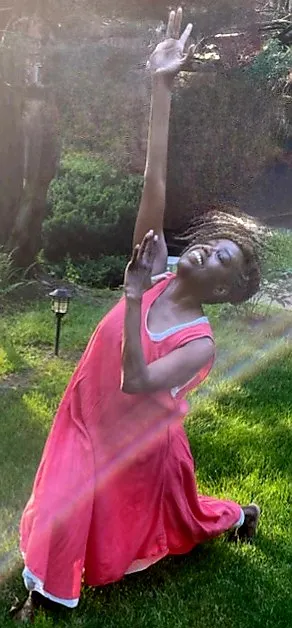 Coach Judy Handwerker performs an expressive dance and movement pose outdoors on a lush green lawn, kneeling with one arm reaching skyward and the other across her chest. Wearing a flowing coral dress, she smiles joyfully as sunlight streams through the scene, creating a radiant effect. The image conveys freedom, praise, vitality, graceful movement, self-expression, and holistic wellness.