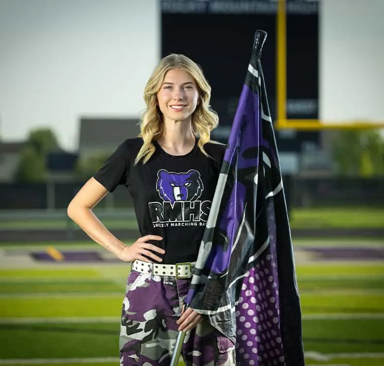 Color guard performer in black RMHS marching band t-shirt with purple bear mascot logo, holding performance flag with purple and black design, standing on football field