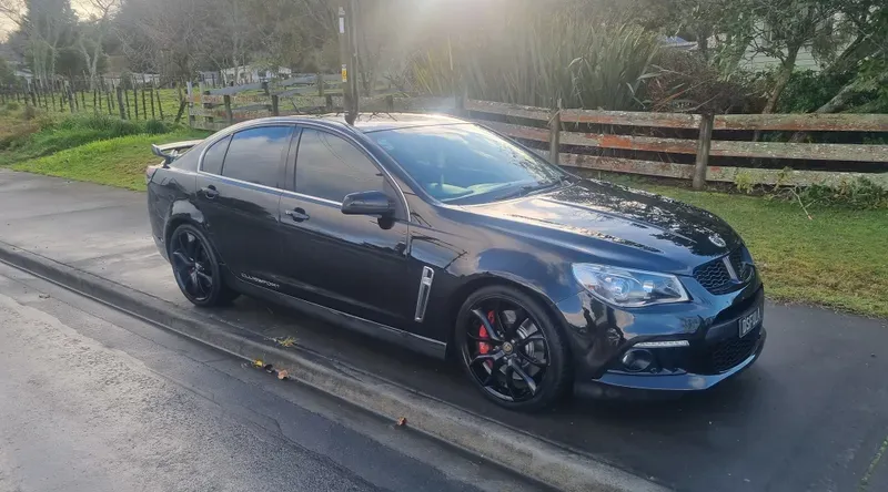 A black Holden Commodore Clubsport sedan is parked on the side of a suburban road on a cloudy day. The car features black alloy wheels with red brake calipers, a rear spoiler, and tinted windows. The background includes a wooden fence, green fields, and trees, with soft sunlight filtering through the clouds.