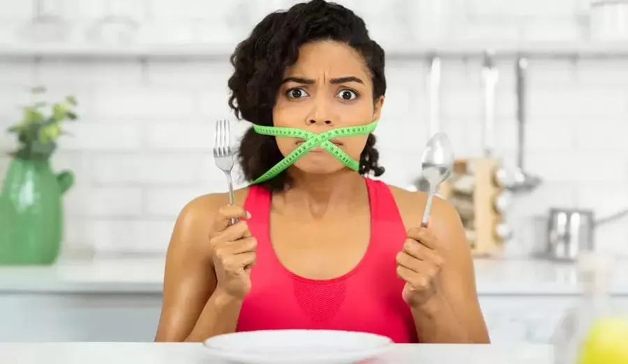 A woman sitting in front of an empty plate holding up a knife and fork with her mouth taped shut with measuring tape. Concept: Fasting 