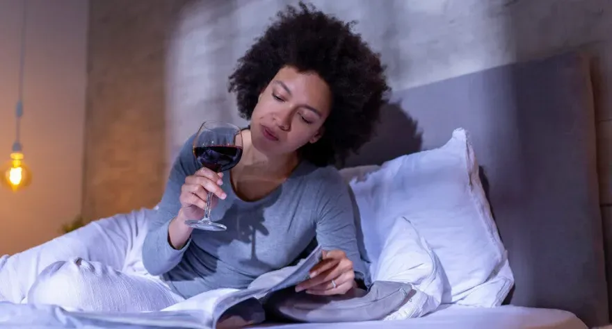 A woman in bed drinking a glass of wine while reading.