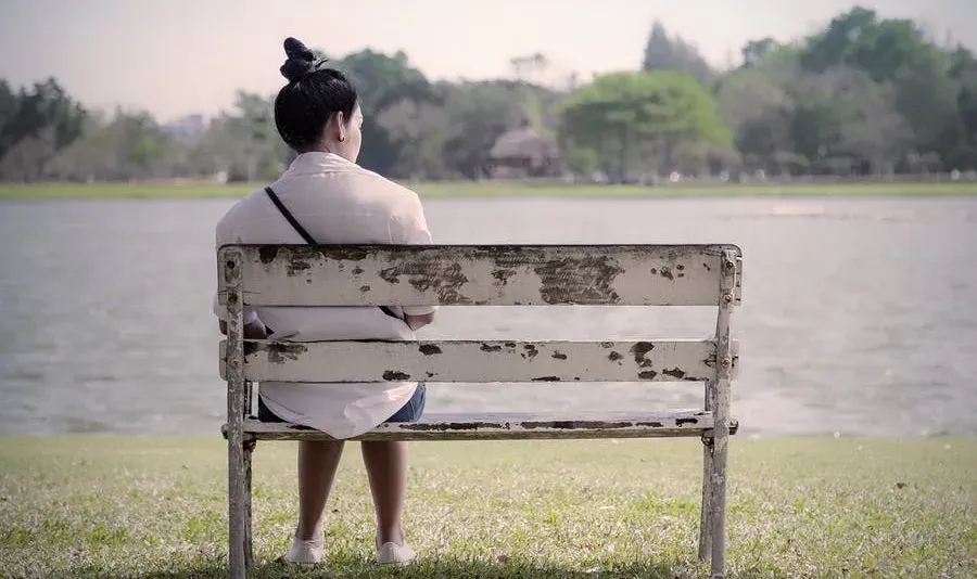 A woman sitting alone by herself on an old, worn-out park bench by a lake.