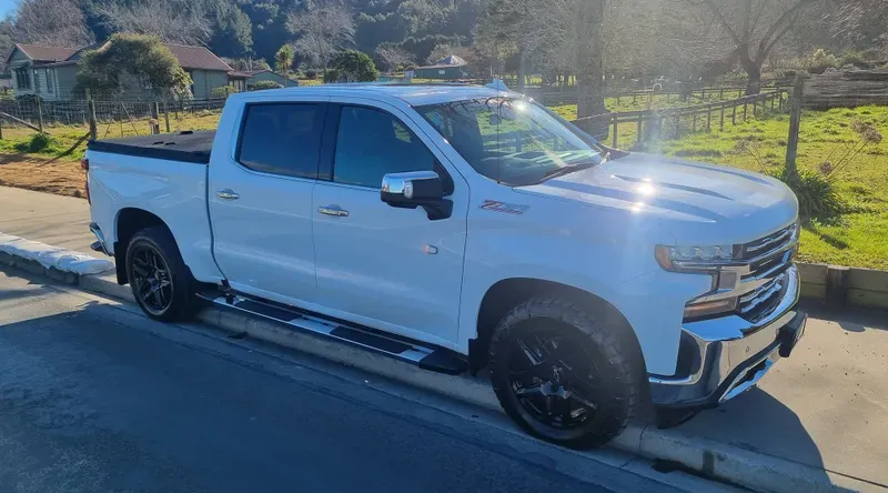 A white Chevrolet Silverado pickup truck is parked on the side of a suburban road on a sunny day. The truck features black alloy wheels, a black bed cover, chrome accents, and tinted windows. The background includes green fields, trees, and a few houses, with clear blue skies and bright sunlight illuminating the scene.