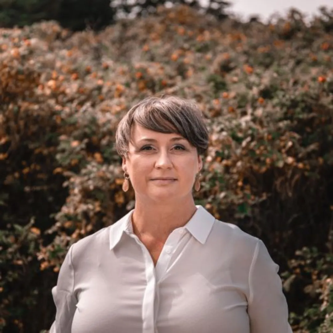 A head shot of Director-Founder, Michelle Hurlburt wearing a white blouse, looking directly at the camera. In the background are brownish-brown bushes with tiny orange flowers. 