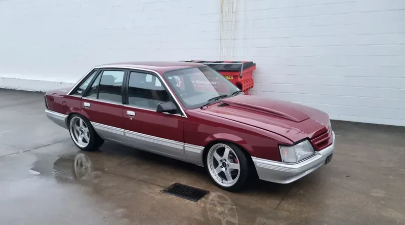 A maroon and silver Holden VK Commodore sedan is parked on a wet concrete surface near a white wall. The car features a custom hood, silver alloy wheels, and a lowered stance. Reflections of the car and surroundings are visible on the wet ground. A red and yellow dumpster is partially visible in the background.