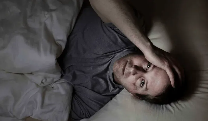 A man awake in his bed in the middle of the night; his hand on head and visibly frustrated.
