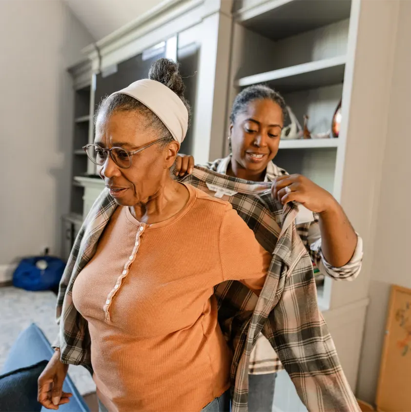 In this image, two women are engaged in a dressing activity in a cozy interior setting. The older woman, wearing a light-orange long-sleeve top and glasses, is in the foreground, appearing focused as she is assisted in putting on a plaid shirt by a younger woman behind her. The younger woman, with her hair tied back, looks cheerful and attentive, helping with the sleeves of the shirt. The background features a neatly arranged living space with light-colored walls, a bookshelf, and a comfortable seating area. The word 