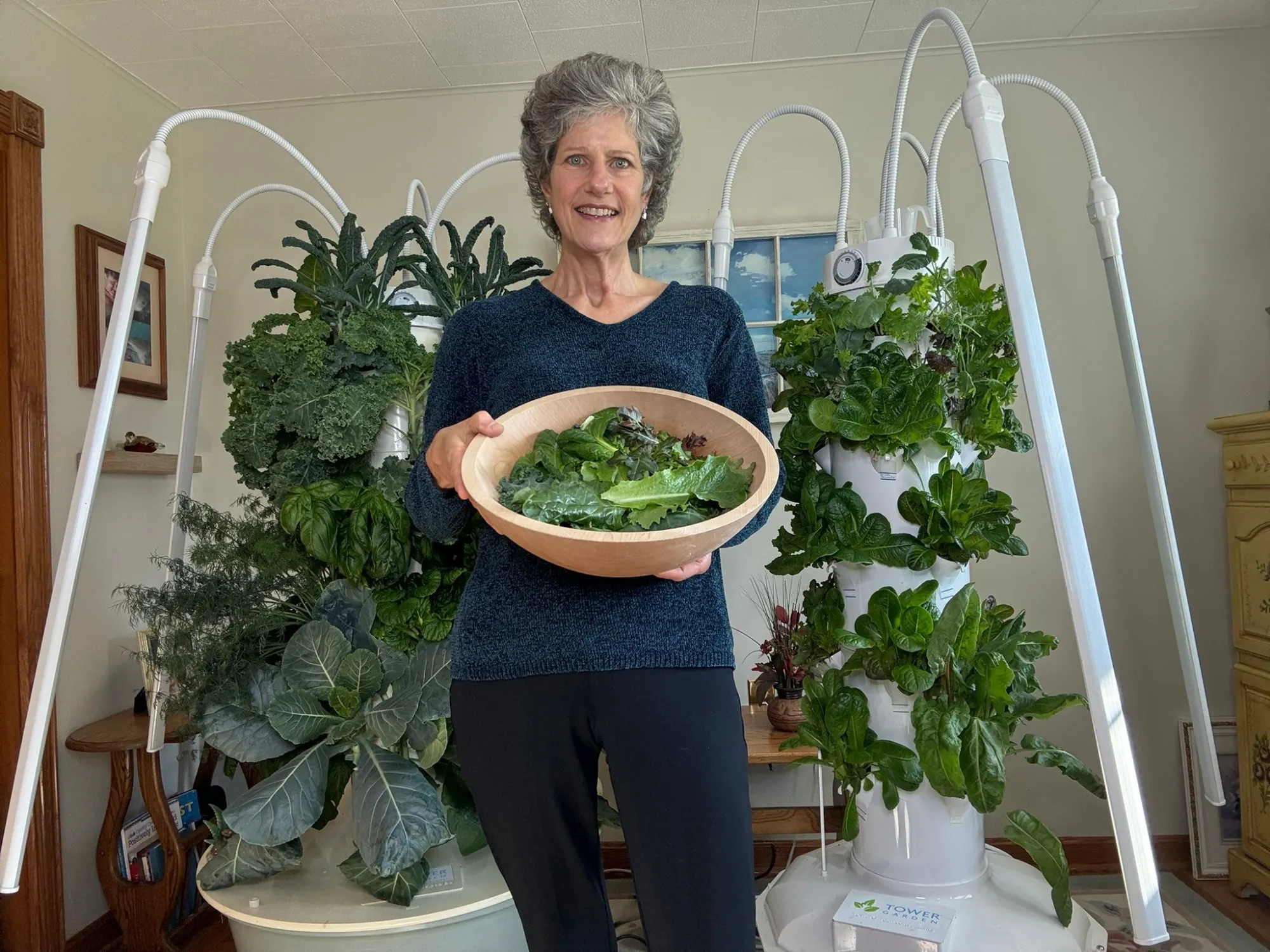 Karen holding a bowl of freshly harvested greens in front of two indoor Tower Garden systems.