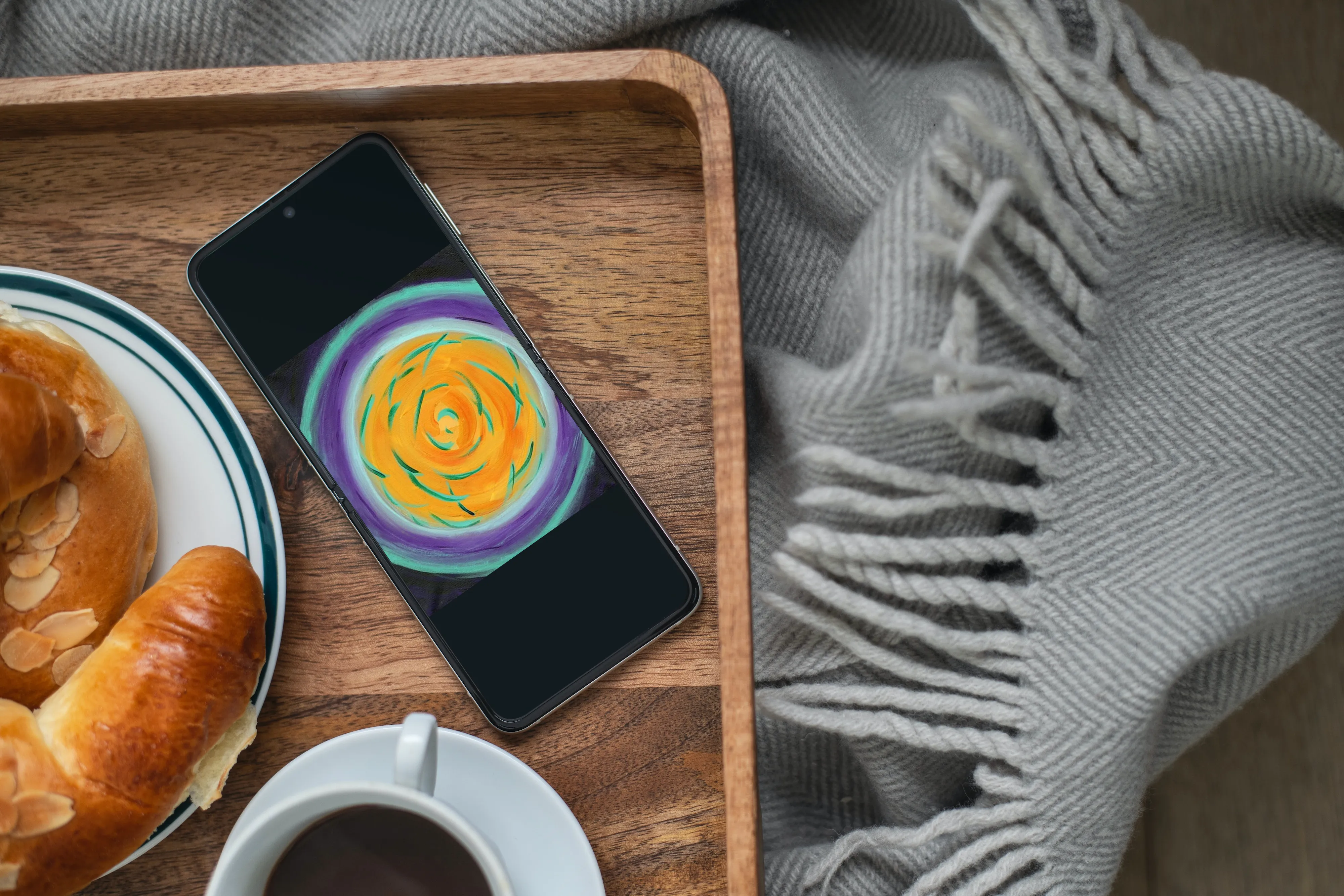 Personal Alignment Painting orange, yellow, teal, indigo  image on smartphone, laying on wooden tray with cup of coffee, croissant, next to gray blanket with tassels.
