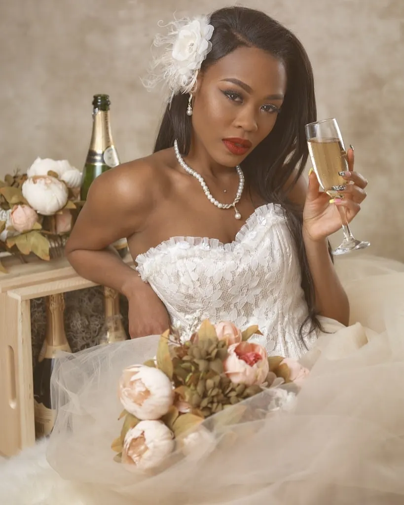 Dark skinned woman posed seated for a bridal portrait wearing a wedding dress, pearls, a flower in her hair and holding a bouquet in one hand with a champagne glass in the other