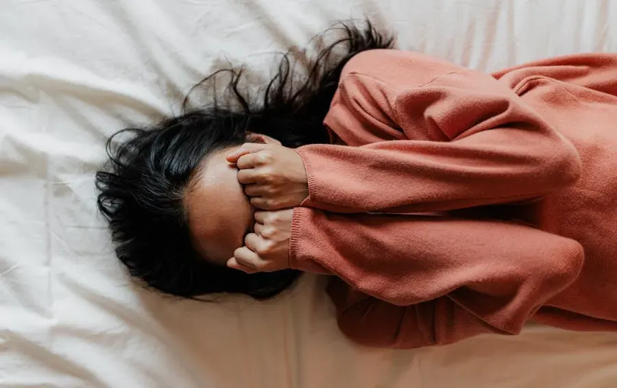 A woman lying on her bed with her hands covering her face appearing to be distressed.