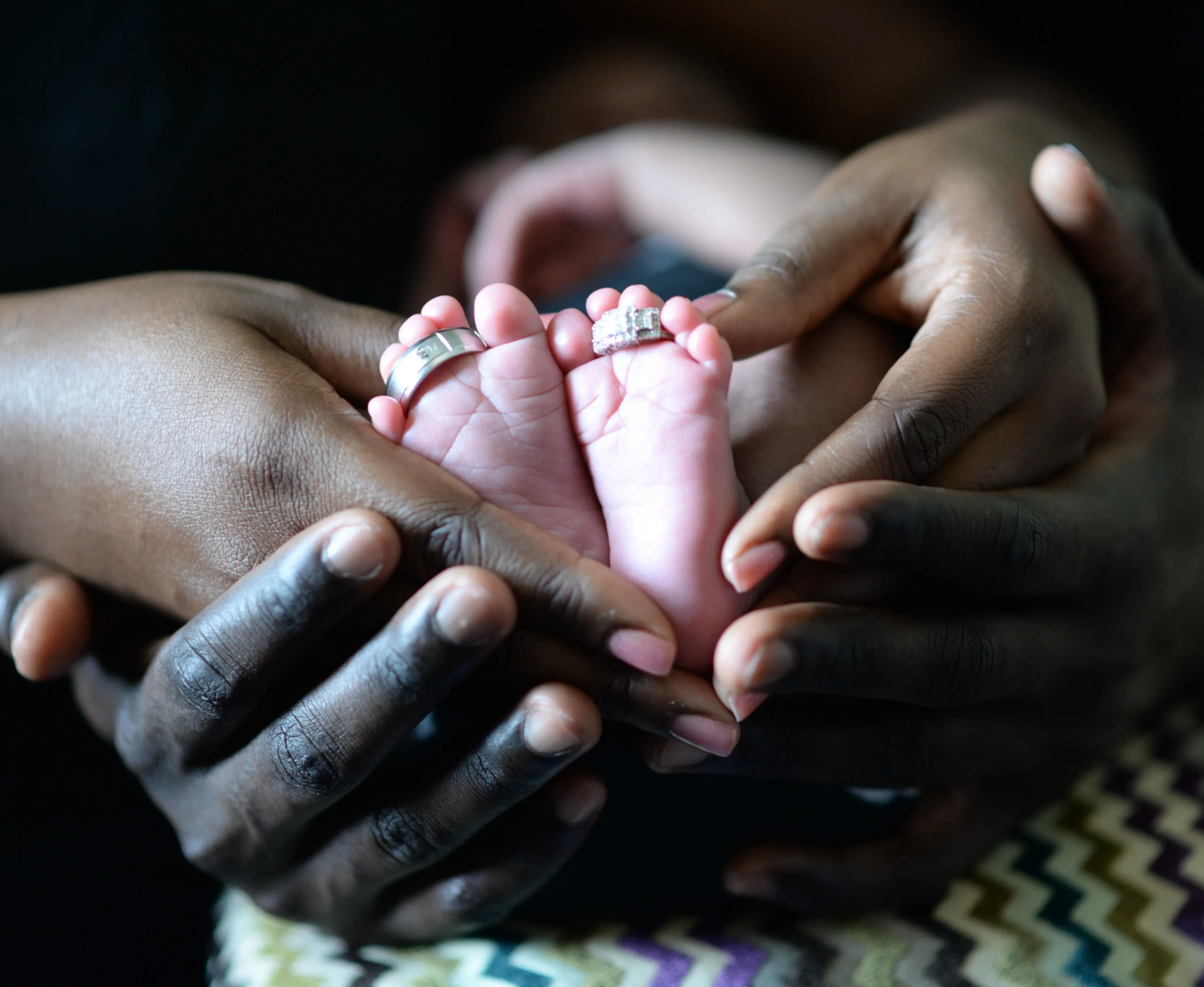 Close up image of parent's wedding rings on newborn's toes while newborn's feet are being held in parents' hands.