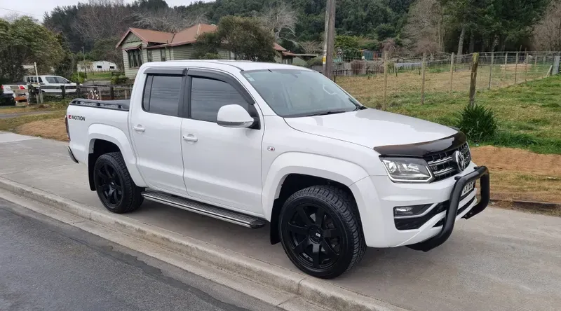 A white Volkswagen Amarok V6 4MOTION pickup truck is parked on the side of a suburban road. The vehicle features black alloy wheels, tinted windows, a black bull bar, and side steps. The background includes a rural setting with green fields, trees, and houses, with overcast skies above.