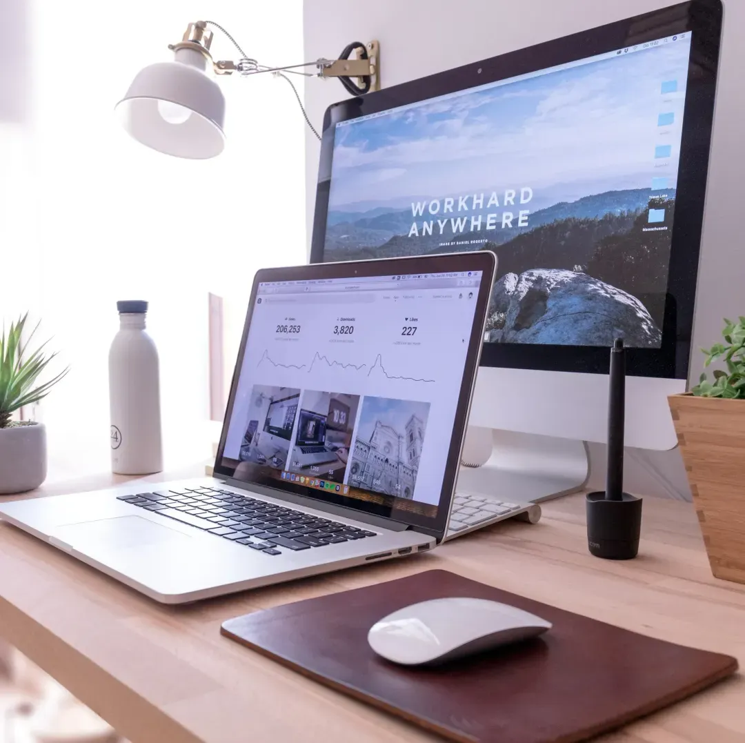 A well-organized workspace with a laptop displaying graphs and analytics on its screen, indicative of remote work or freelancing. Behind it, a desktop monitor with 'WORK HARD ANYWHERE' as the wallpaper, symbolizes the flexibility of modern work. The desk is accessorized with a white water bottle, a green plant, a desk lamp, a stylus, and a leather notebook, creating a productive and stylish home office environment.