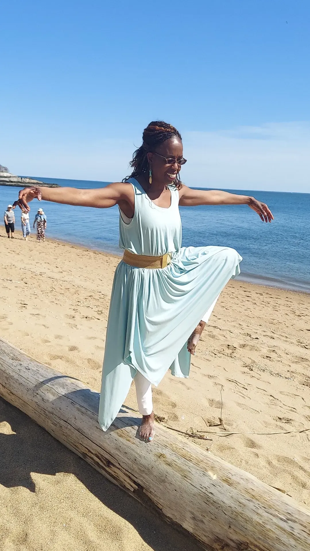 Coach Judy Handwerker balances gracefully on a driftwood log along a sandy beach shoreline, performing a mindful movement pose with arms extended and one knee raised. Dressed in a flowing light blue outfit, she overlooks the water under a clear sky. The image reflects balance, vitality, wellness, movement, confidence, and the connection between physical activity and inner wellbeing.