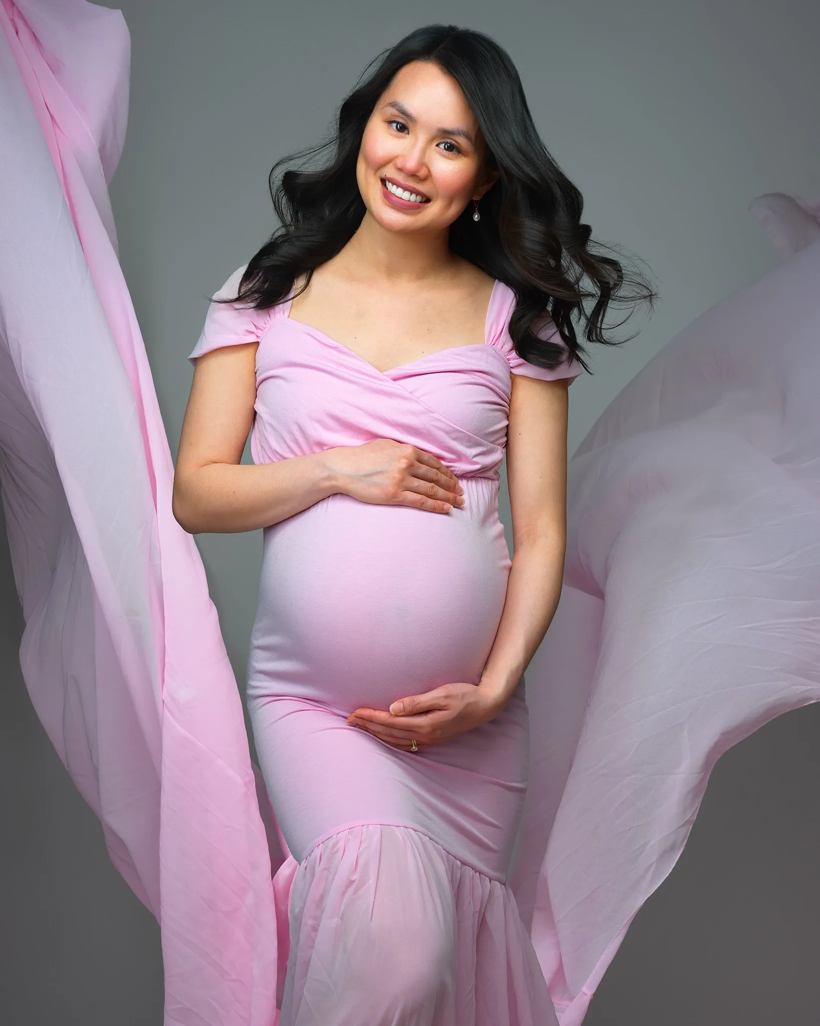 Maternity portrait of asian woman in pink flowing dress posed in front of a grey backdrop