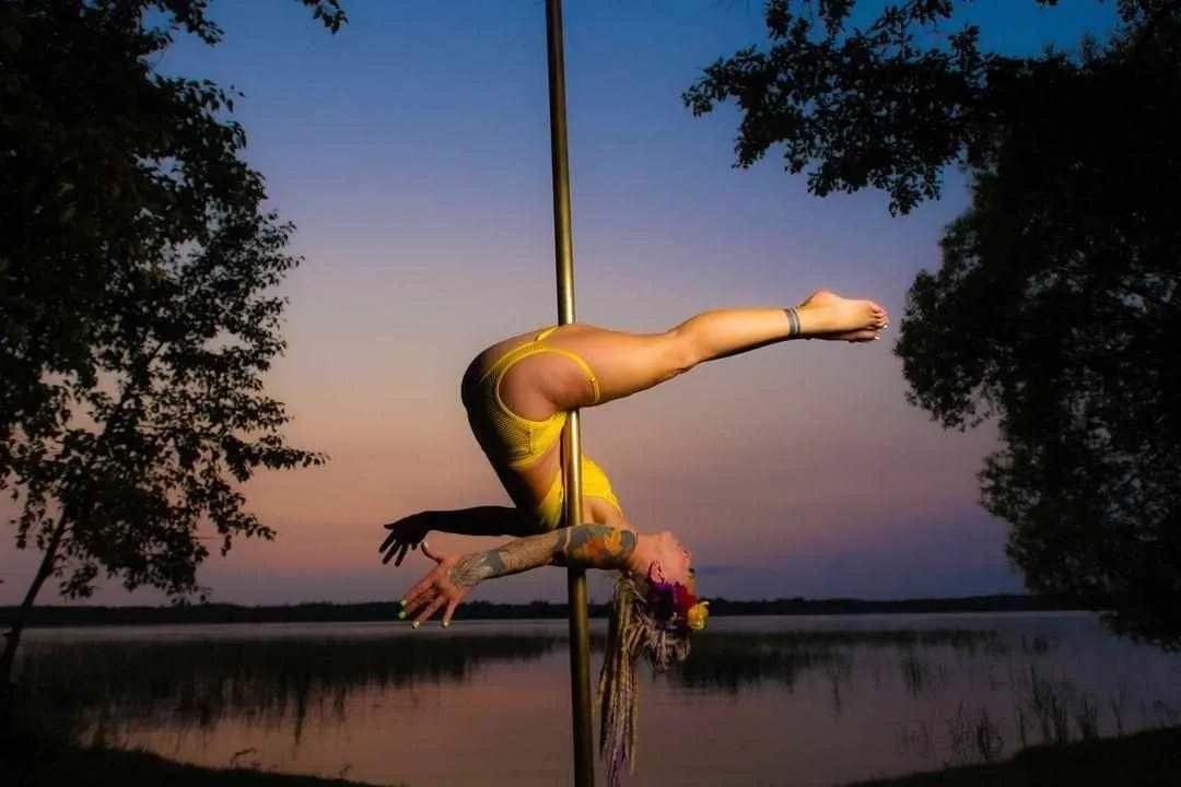 Amanda is inverted in a hip hold on the pole in front of a sunset and lake. 