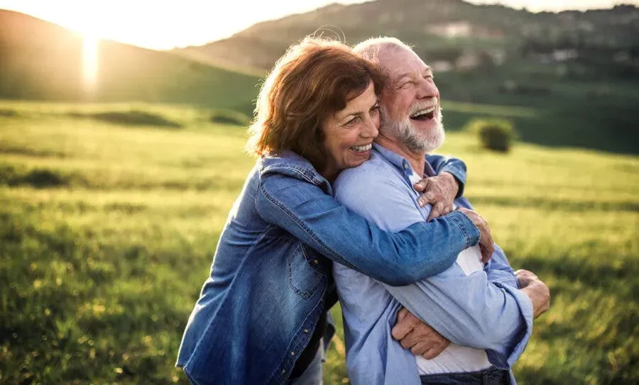 an elderly couple in a field, embracing and laughing together with a sunset in the background.