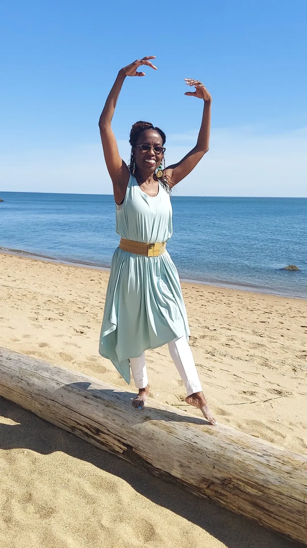 Coach Judy Handwerker balances gracefully on a driftwood log along a sandy beach, posing with raised arms in a ballet-inspired movement. Wearing a flowing light blue dress with white leggings, she smiles while overlooking calm ocean waters beneath a clear blue sky. The image symbolizes balance, joy, praise, mindful movement, wellness, and active aging.