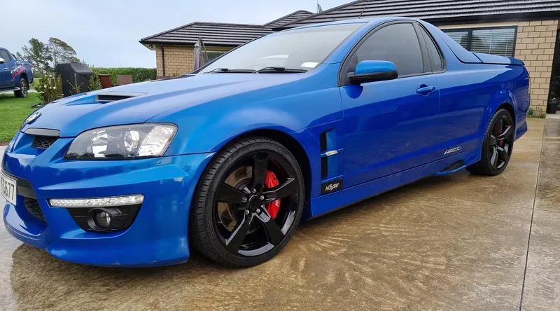  blue HSV Maloo ute is parked on a wet driveway in front of a brick house. The vehicle features black alloy wheels with red brake calipers, a hood scoop, and aggressive styling elements. The glossy finish reflects the overcast sky, creating a vibrant contrast with the muted surroundings. In the background, another blue vehicle and a well-maintained lawn are visible.