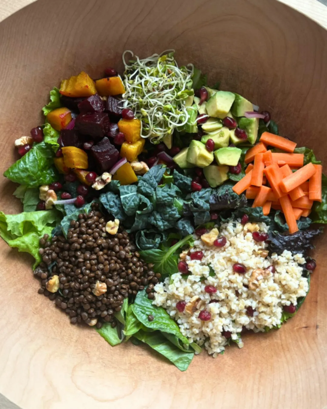 Winter Jewel Salad of the Week in a wooden bowl with leafy greens, kale, lentils, bulgur, roasted beets, avocado, carrots, pomegranate seeds, and sprouts.