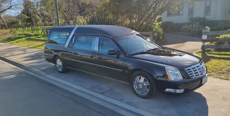 A black Cadillac hearse is parked on the side of a suburban road on a sunny day. The vehicle features chrome accents, tinted windows, and a vinyl roof. In the background, there are green fields, trees, and houses, with clear skies and bright sunlight illuminating the scene. The hearse has a classic and elegant design, suited for its solemn purpose.