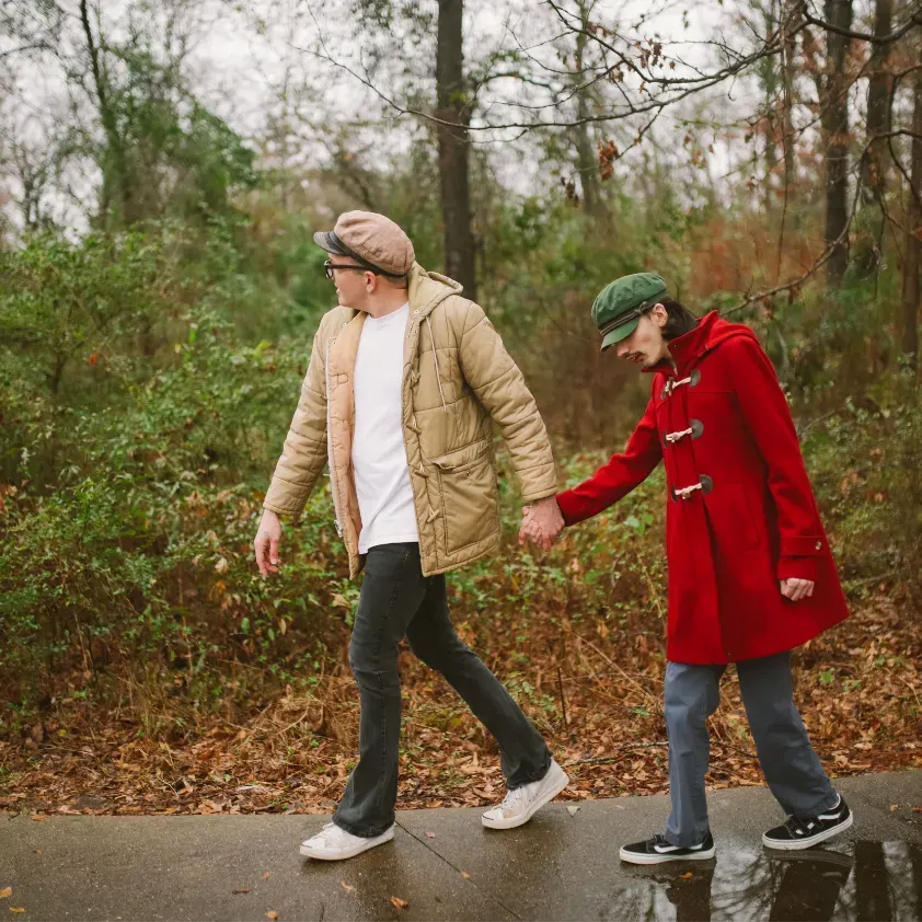 A serene outdoor scene shows a woman trailing behind a man, having difficulty keeping up. The woman wears a light tan puff jacket, white t-shirt, and black pants, while the man is dressed in a vibrant red coat and blue jeans. Both wear stylish hats, with the woman in a beige cap and the man in a green one. The word 'WALKING' is displayed at the top, emphasizing the activity. The background features lush greenery and a wet pathway, suggesting recent rainfall.