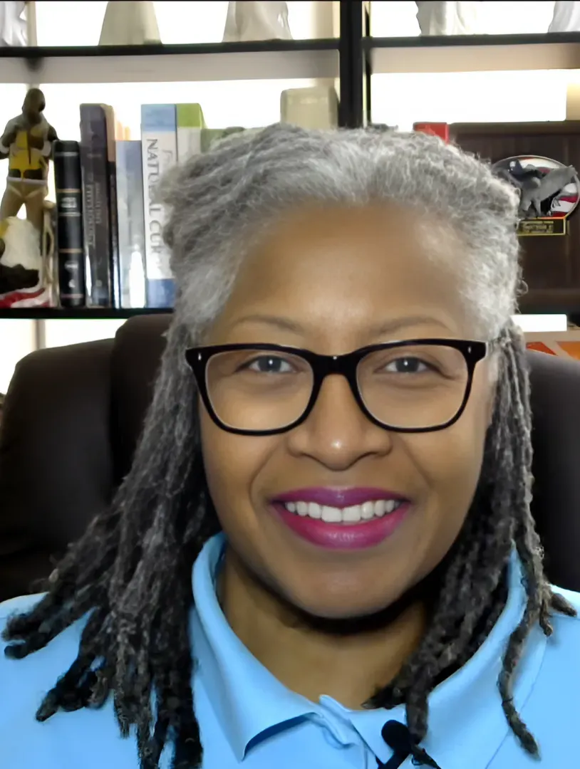 Dr. Lisa smiling, wearing black-rimmed glasses and a light blue shirt, seated in front of a bookshelf with various books and decor. She has gray hair styled in natural locs. The background includes books and small sculptures, suggesting a professional, intellectual setting.