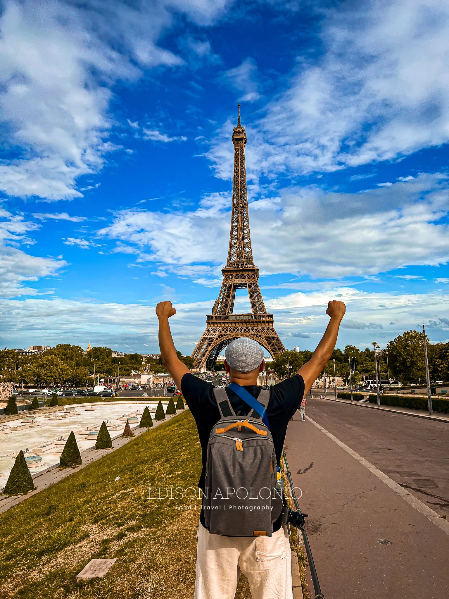 man wearing a black shirt and a pair of white or cream pants plus a grey cap on, carrying a grey backpack strapped to his back, standing with both hands closed fist, both arms are held up to the sky on a Victory pose, facing The Eiffel Tower in the distance.