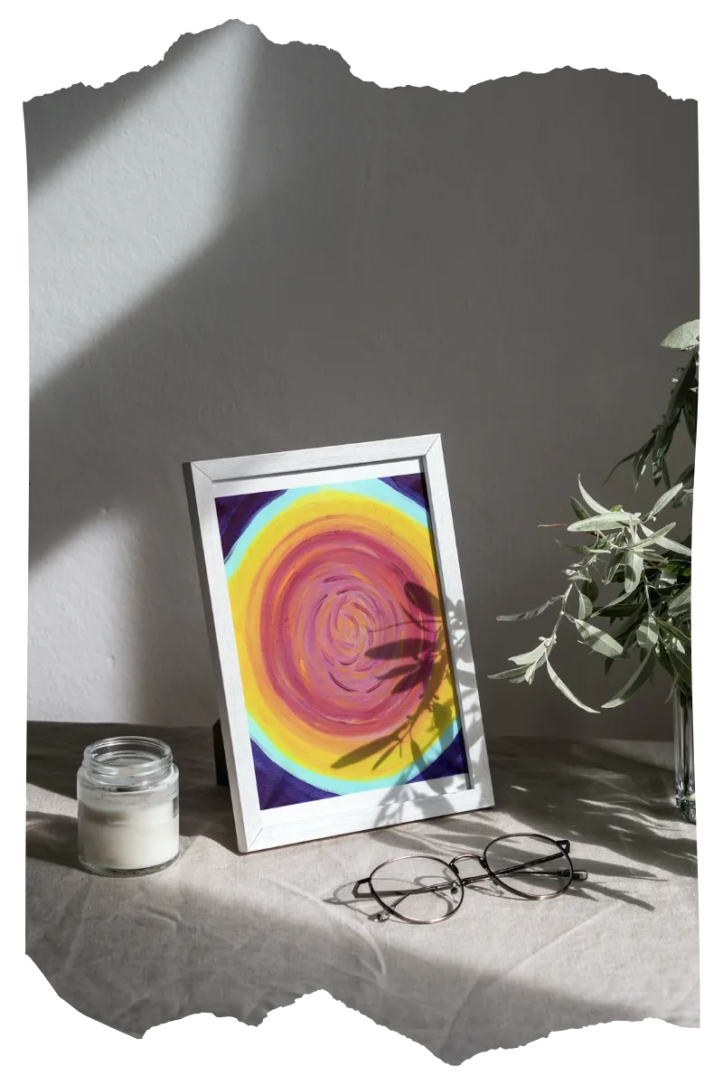 Graytone shadows on wall and desk with plant, eyeglasses, and tabletop framed Personal Alignment Painting in pinks, yellows, oranges, sky blue, and indigo.