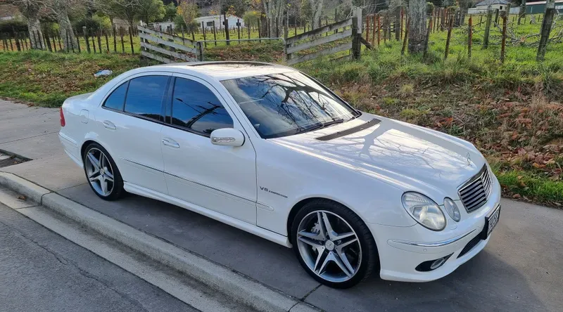 A white Mercedes-Benz E55 AMG sedan is parked on the side of a suburban road. The car features silver alloy wheels, tinted windows, and V8 Kompressor badging. In the background, there are green fields, trees, and a wooden fence, with fallen leaves scattered on the ground. The overcast sky adds a calm, autumnal atmosphere to the scene.