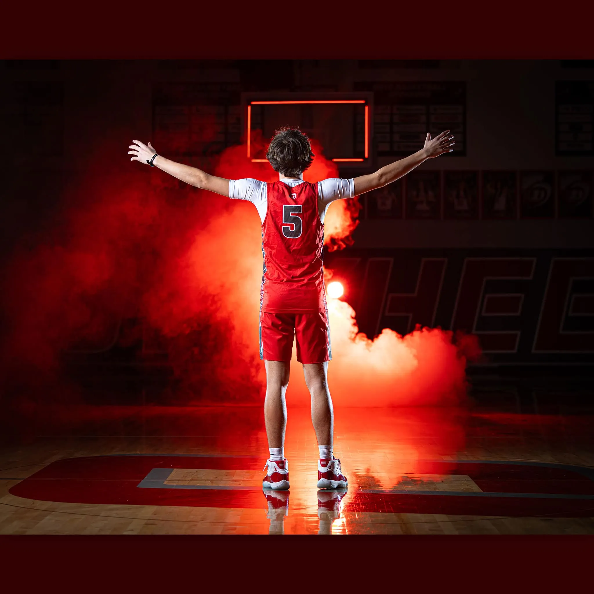 Basketball player in red jersey number 5 stands with arms spread wide on a basketball court, backlit dramatically with red smoke effects surrounding them