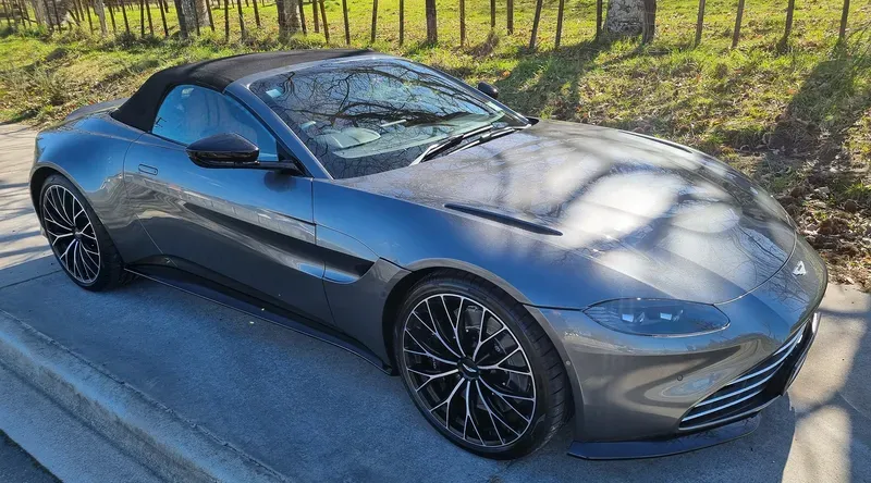 A sleek grey Aston Martin Vantage convertible is parked on the side of a suburban road. The car features a black soft-top roof, black multi-spoke alloy wheels, and a low, aerodynamic design. Sunlight filters through the trees, casting shadows on the car's shiny surface. The background includes a grassy area with trees and a wooden fence under clear skies.