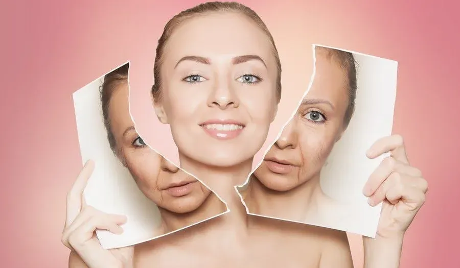 A young woman with clear, youthful skin smiling brightly. She is holding up a torn photograph that was previously covering her face. The photograph depicts an older version of herself, with visible wrinkles and signs of aging.