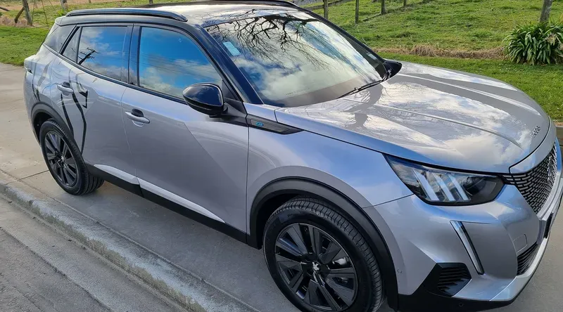 A silver Peugeot 2008 SUV is parked on the side of a suburban road. The vehicle features black alloy wheels, a sleek design with sharp lines, and tinted windows. Reflections of the sky and surrounding trees are visible on the car's shiny surface. The background includes green fields, a fence, and some houses, creating a peaceful rural setting.