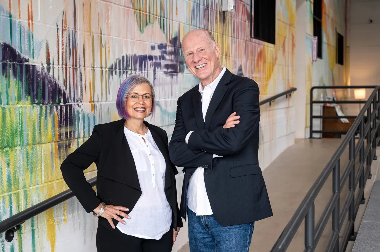 Cora and Steve, CBArt Studios owners, standing together in front of a colorful painted wall