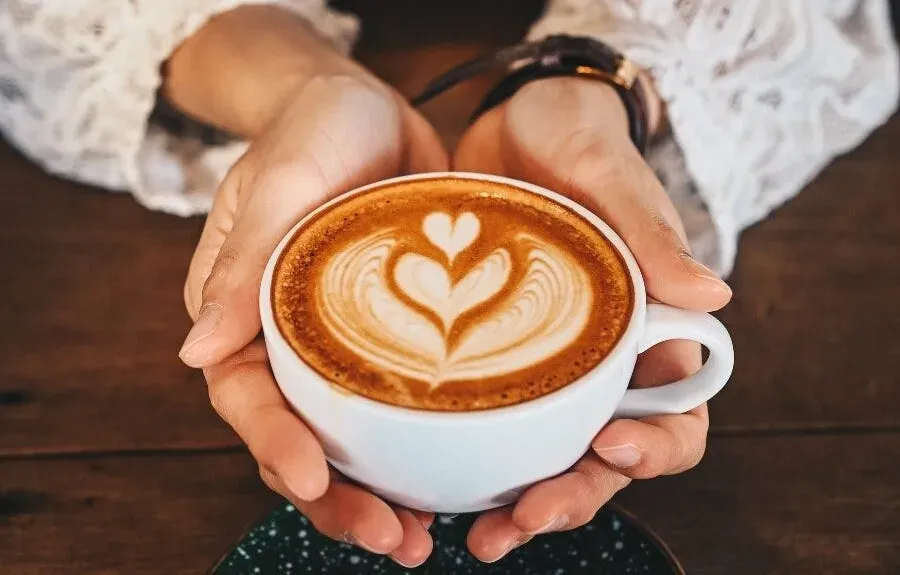 a person holding a cup of coffee with intricate latte art in the shape of a heart, in a warm and cozy setting. 