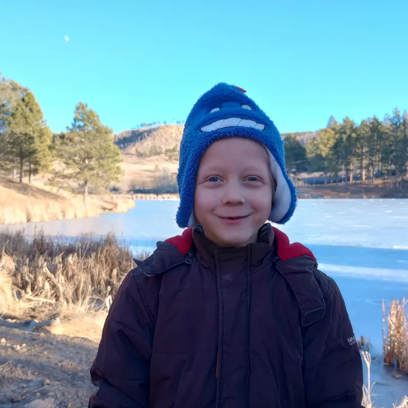 little boy with a smile wearing warm coat, hat standing outside by a pond