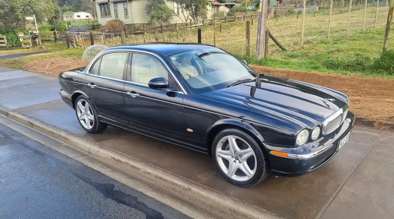 A black Jaguar XJ sedan is parked on the side of a suburban road on a cloudy day. The car features silver alloy wheels, chrome accents, and a classic, elegant design. The background includes green fields, trees, and houses, with soft light reflecting off the vehicle's surface, creating a serene atmosphere.