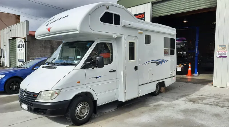 A white Mercedes-Benz Sprinter camper van is parked outside a garage with its door partially open. The vehicle features a raised roof with a sleeping area, blue wave decals on the sides, and a side door. The camper van is parked on a concrete surface, with a blue car and a building labeled 