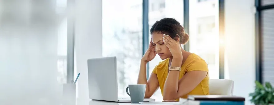 A stressed out looking woman sitting in front of her computer with her eyes closed and rubbing her temples.