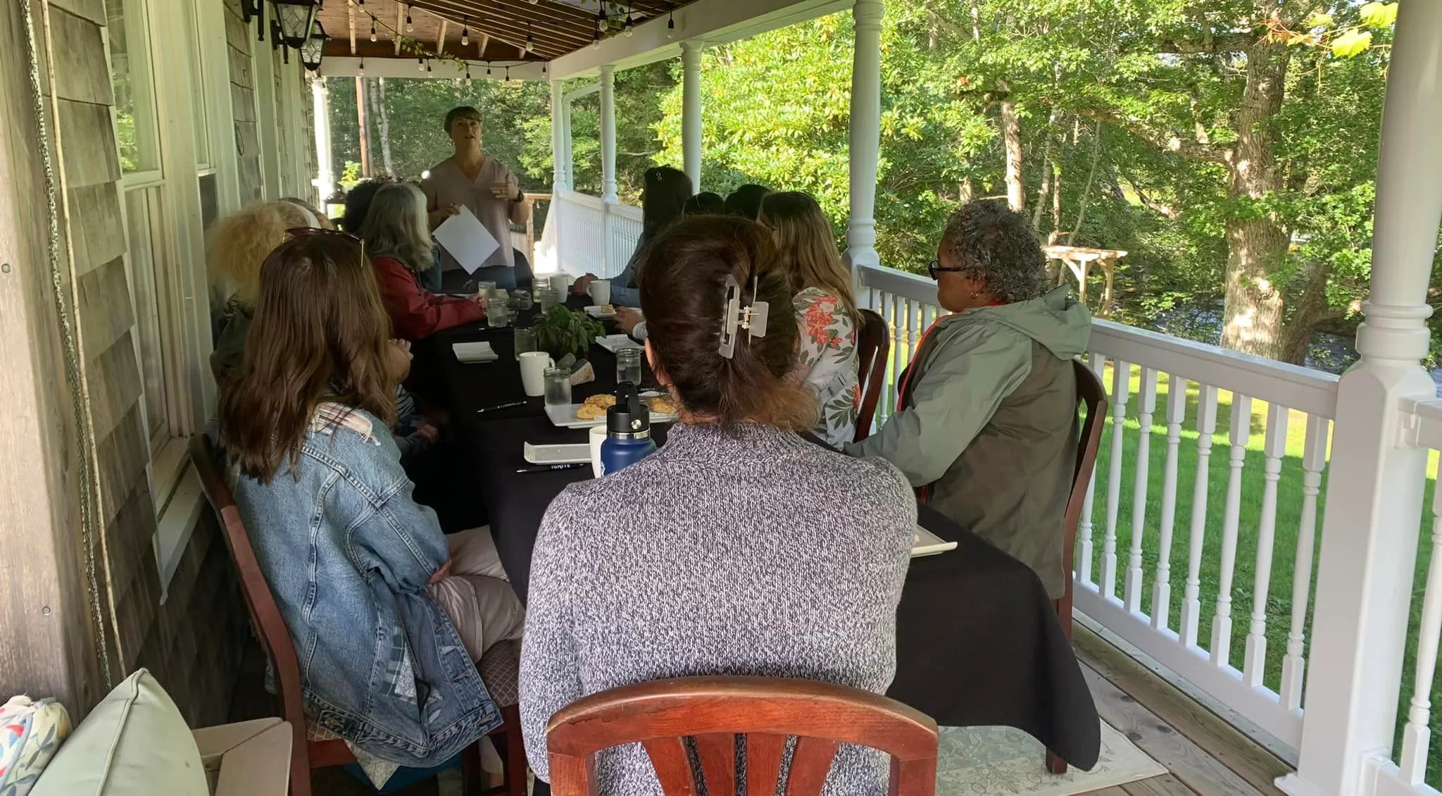 Director-Founder, Michelle Hurlburt standing at the end of a long table on a covered deck. A group women leaders and managers are sitting at the table back-to the camera, listening to Michelle. In the background are trees and green leaves. 