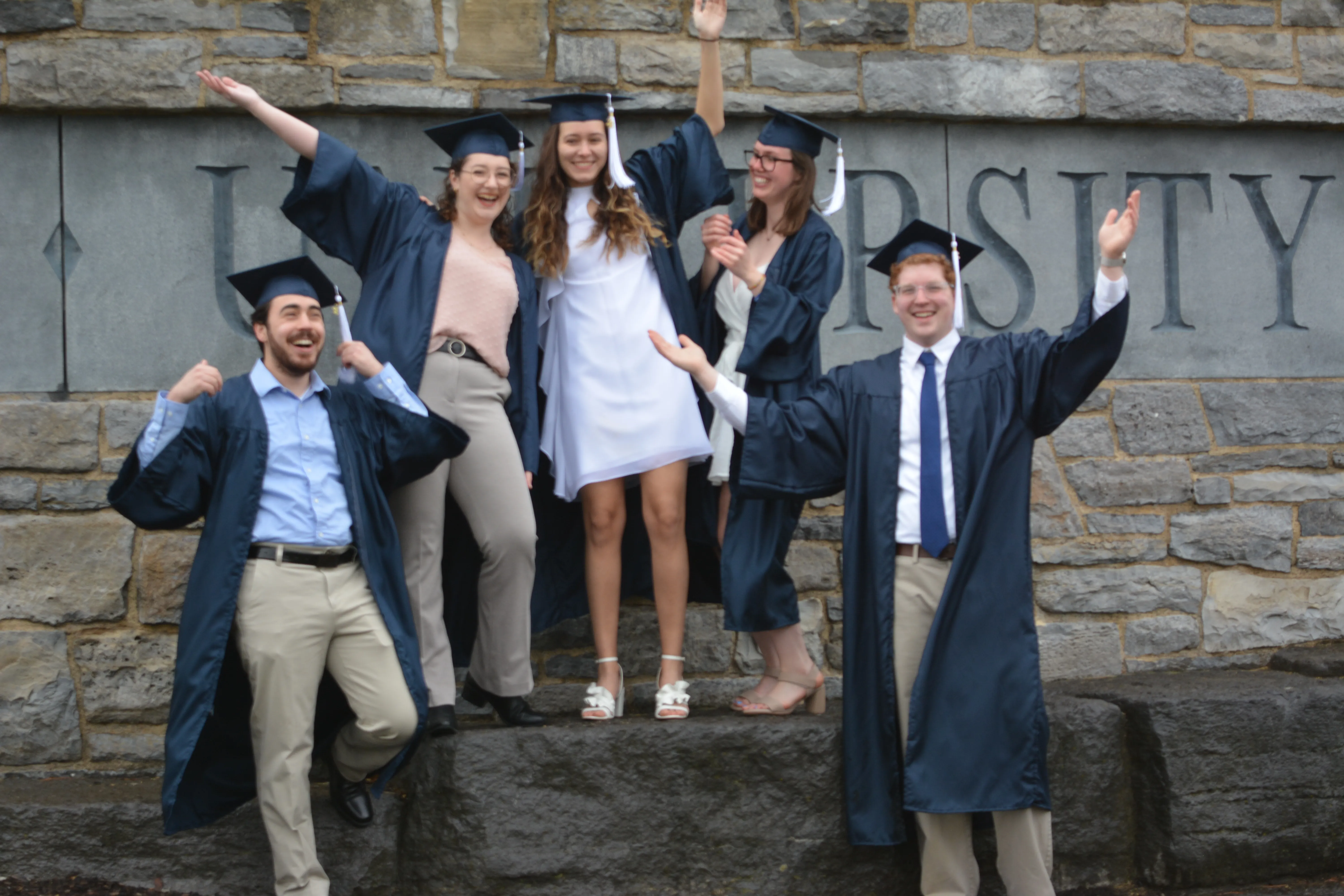 Three young women and two young men wearing blue graduation caps and gowns. All have arms pumped in the air and are smiling